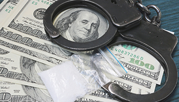 A pair of handcuffs and a bag of crack cocaine are spread out on top of a pile of US dollar bills.